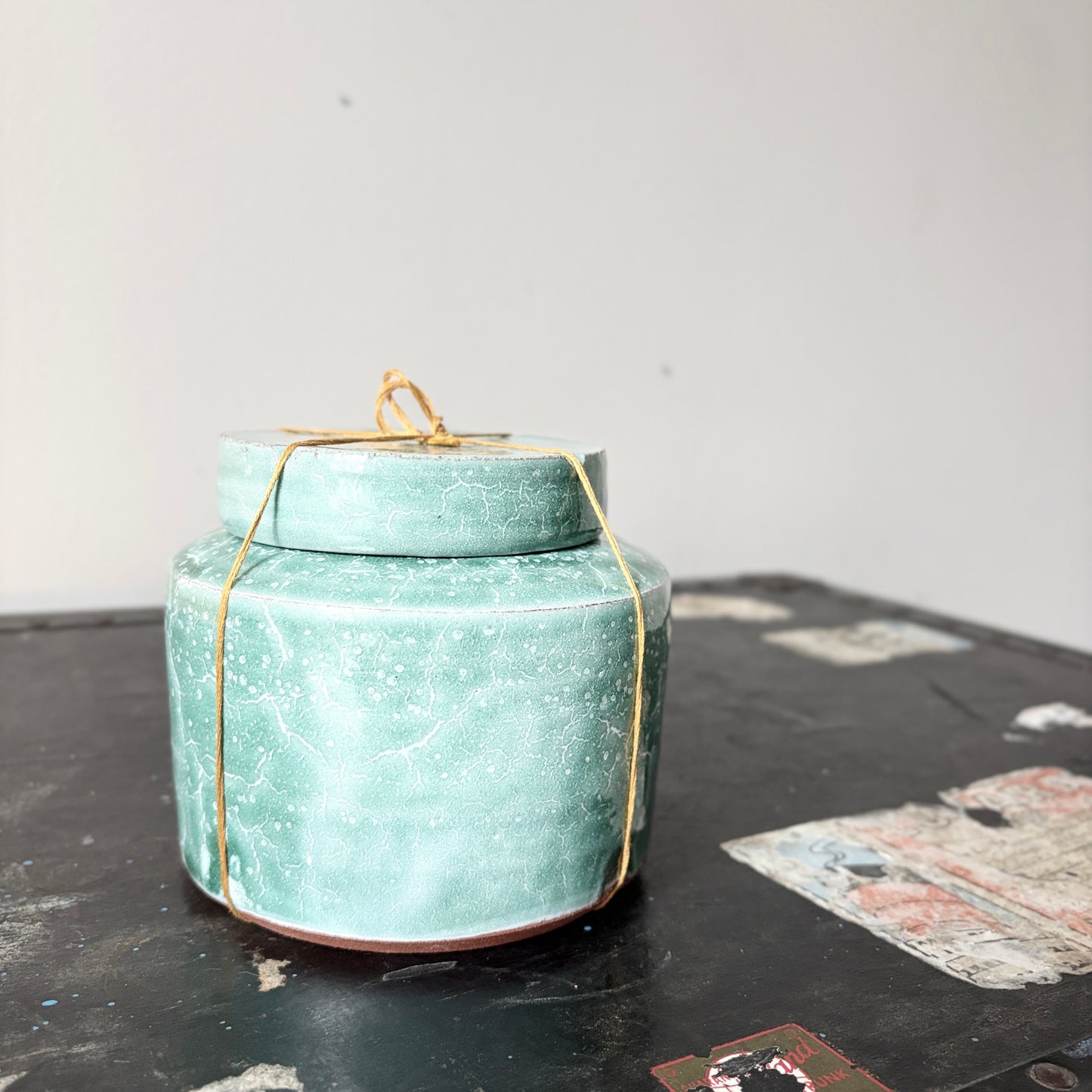 A small turquoise ceramic lidded jar with a golden thread tied around it.