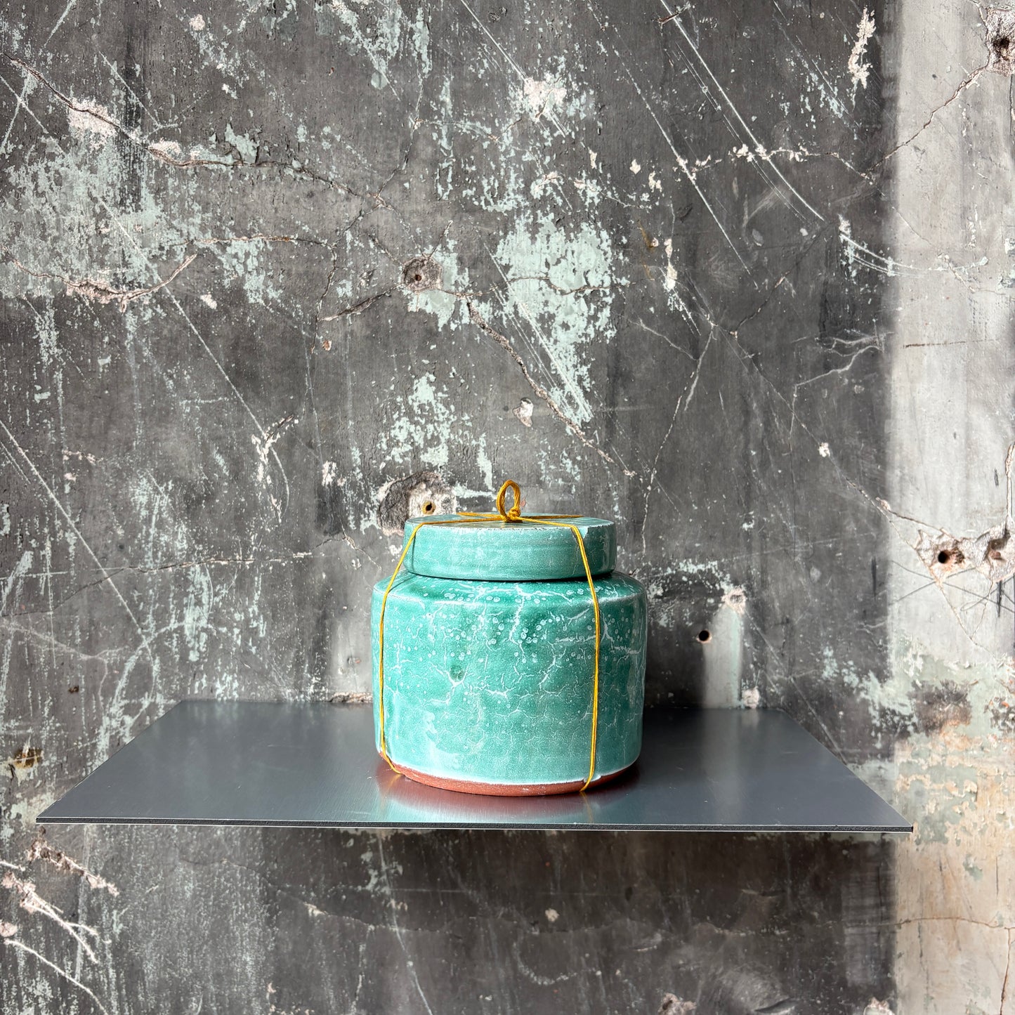 A small turquoise ceramic lidded jar with a golden thread tied around it, displayed against a concrete wall.

