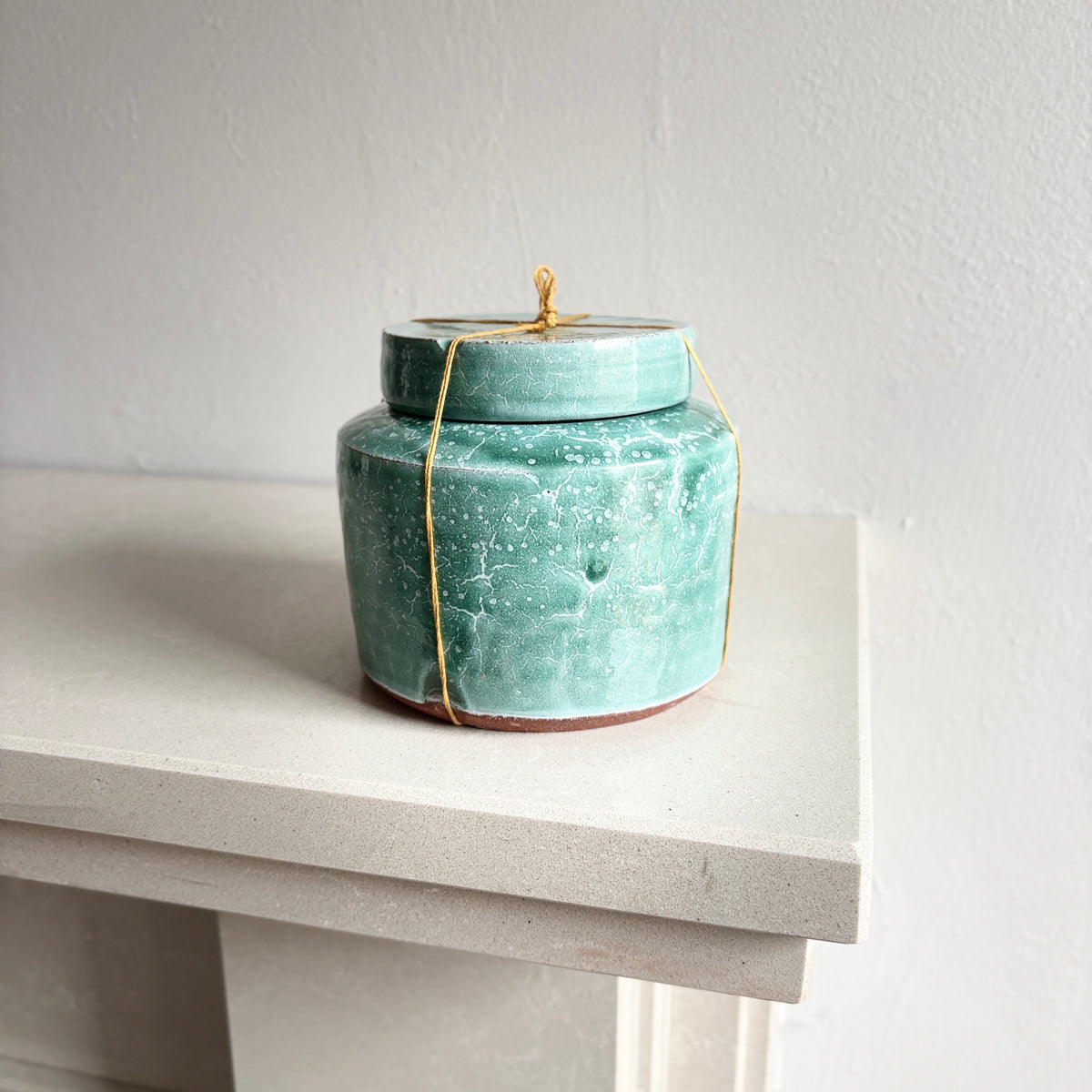 A small turquoise ceramic lidded jar with a golden thread tied around it.